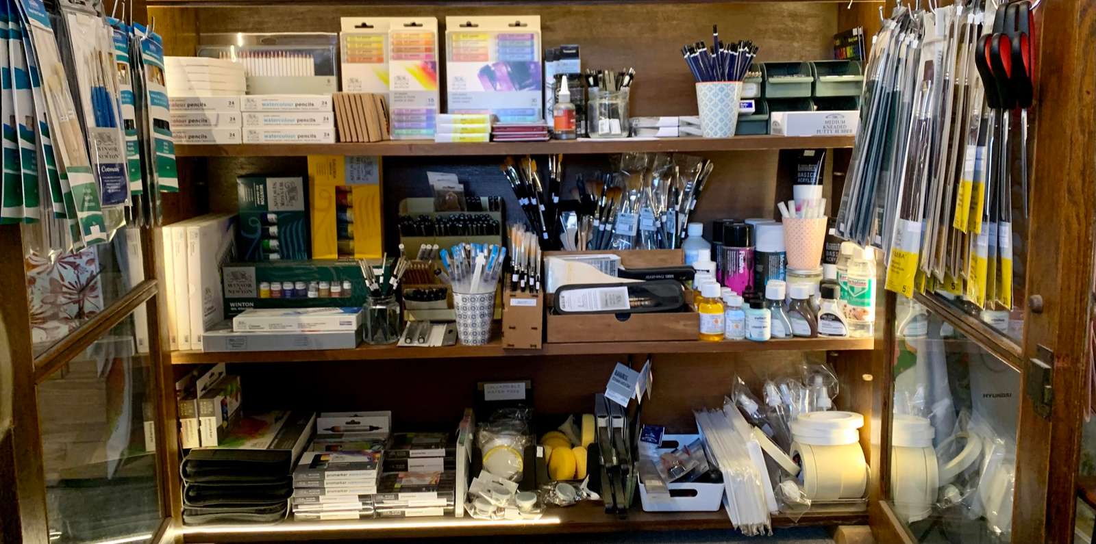 The art room cabinet: shelves of brushes, paint sets, pens, mediums and palettes, with brush packs hanging at either side