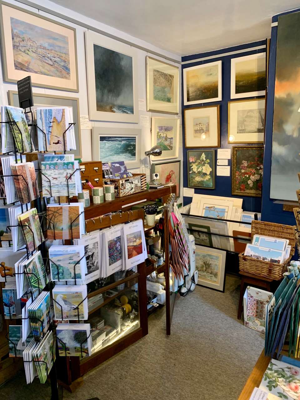 A corner of the gallery: framed paintings hung on white and deep blue walls, with spinners of cards and calendars in front