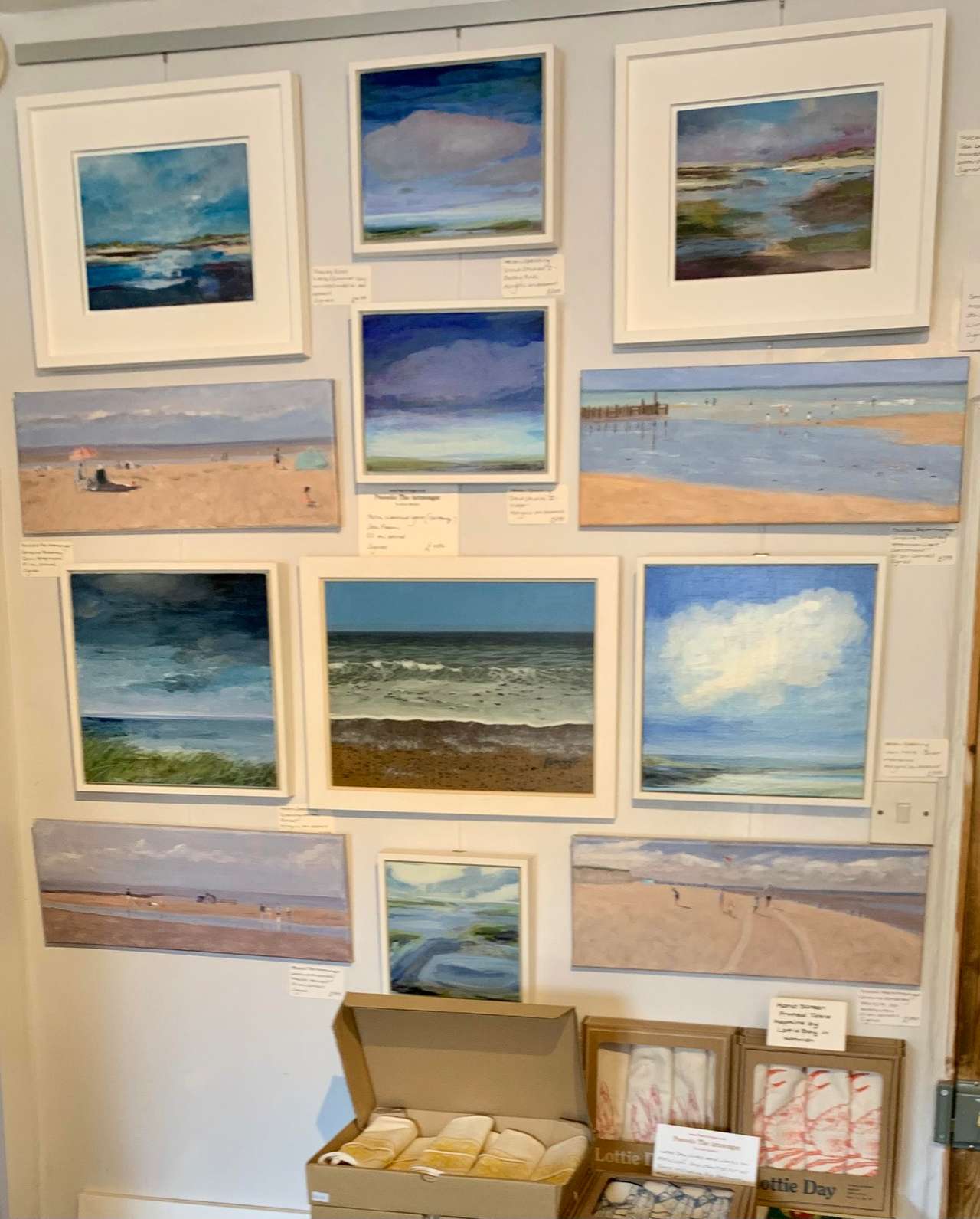 A wall hung with small framed paintings of Norfolk beaches, sea and sky